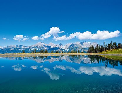 Sicht vom klaren Astbergsee auf eine Bergkulisse und strahlend blauem Himmel.