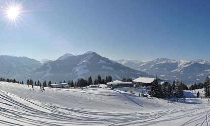 Verschneite Winterlandschaft mit Pisten, Berghütten, Skifahrern und strahlender Sonne über dem Skigebiet Wilder Kaiser – Brixental